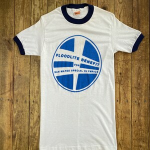 Pu&ograve; includere: Maglietta bianca con un grafico circolare blu e bianco che dice "Floodlite Benefit for San Mateo Special Olympics". La maglietta ha un bordino blu scuro.
