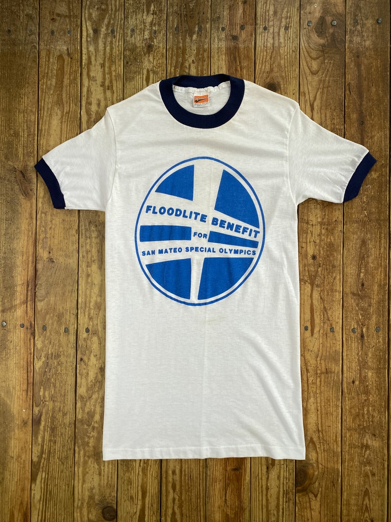 Pu&ograve; includere: Maglietta bianca con un grafico circolare blu e bianco che dice "Floodlite Benefit for San Mateo Special Olympics". La maglietta ha un bordino blu scuro.