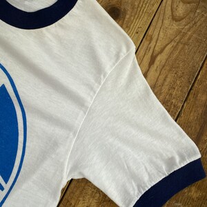 Pu&ograve; includere: Maglietta bianca a maniche corte con un design grafico blu e bianco. La maglietta ha un collo e polsini a costine blu scuro.