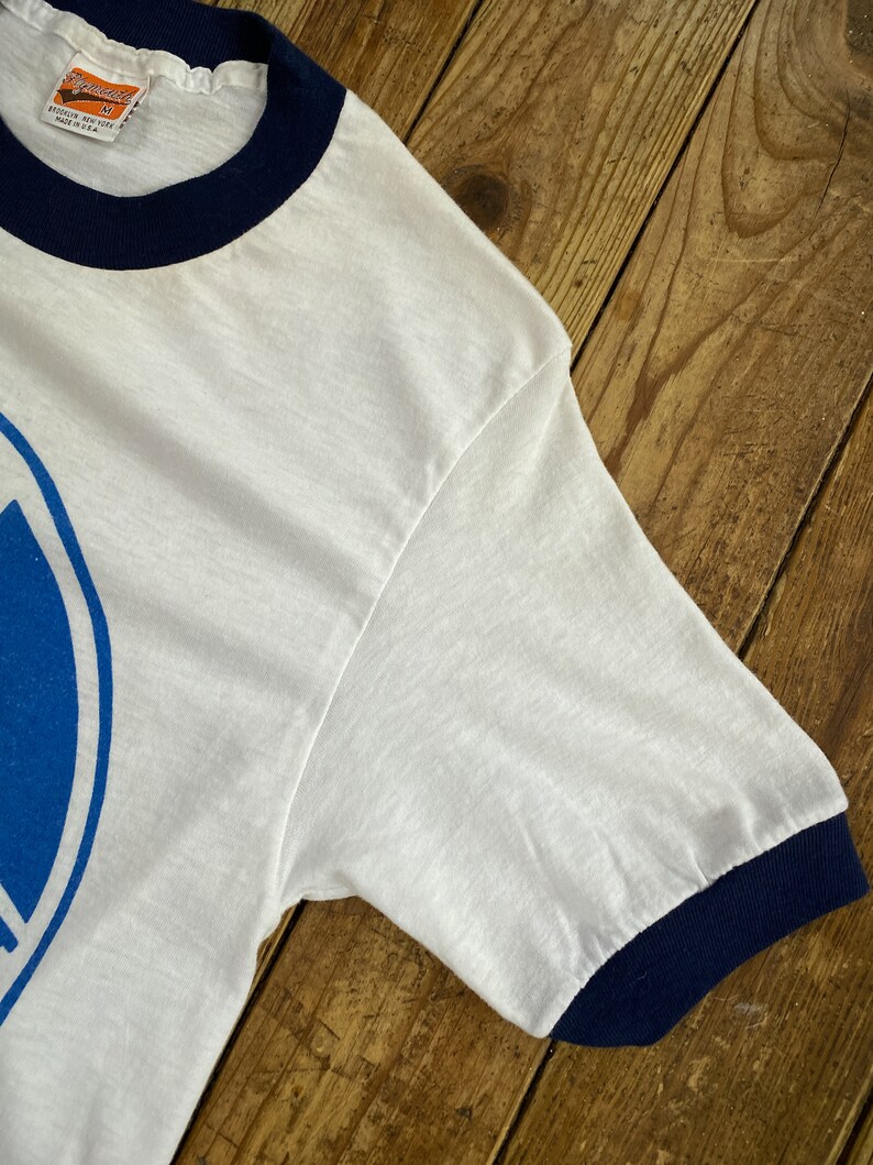 Pu&ograve; includere: Maglietta bianca a maniche corte con un design grafico blu e bianco. La maglietta ha un collo e polsini a costine blu scuro.