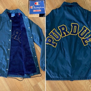 May include: A navy blue Champion jacket with a Purdue logo on the back. The jacket has a snap closure and a drawstring at the bottom. The inside of the jacket is lined with a blue fleece material.