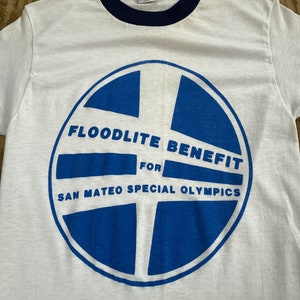 Pu&ograve; includere: Maglietta bianca con collo a righe blu e un grafico circolare blu con il testo "Floodlite Benefit for San Mateo Special Olympics".