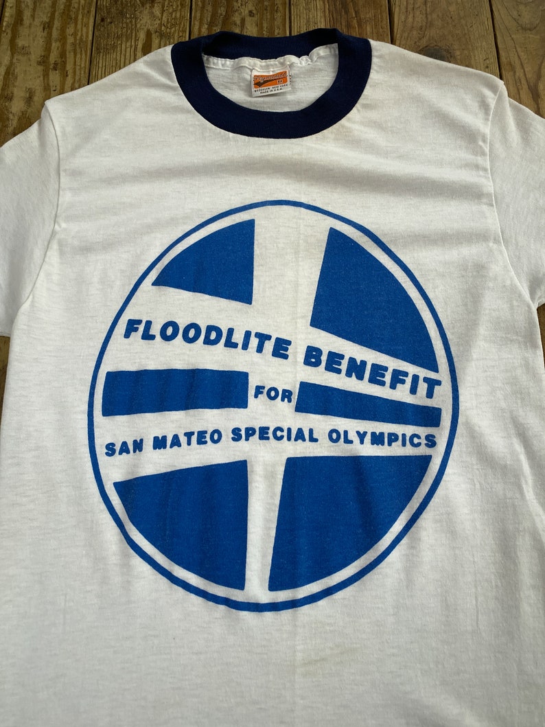 Pu&ograve; includere: Maglietta bianca con collo a righe blu e un grafico circolare blu con il testo "Floodlite Benefit for San Mateo Special Olympics".