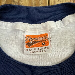 Pu&ograve; includere: Maglietta bianca con collo a costine blu navy e un'etichetta che recita "Plymouth" in lettere marroni su sfondo arancione. L'etichetta riporta anche "M", "Brooklyn New York" e "Made in U.S.A."