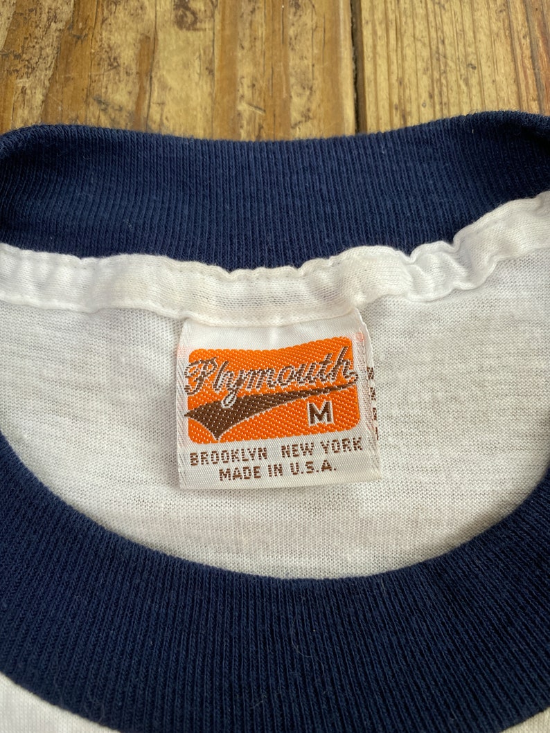 Pu&ograve; includere: Maglietta bianca con collo a costine blu navy e un'etichetta che recita "Plymouth" in lettere marroni su sfondo arancione. L'etichetta riporta anche "M", "Brooklyn New York" e "Made in U.S.A."