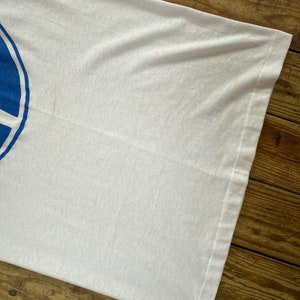 Pu&ograve; includere: Una maglietta bianca con un design circolare blu. La maglietta &egrave; piegata e appoggiata su una superficie di legno.