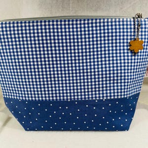 May include: A blue and white gingham patterned cosmetic bag with a navy blue polka dot bottom. The bag has a silver zipper and a wooden flower charm attached. The bag is designed to hold makeup and other beauty products.