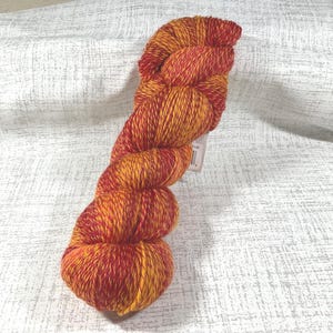 May include: A skein of hand-dyed yarn in shades of red and yellow. The yarn is tightly wound, showing a blend of colors. The texture appears soft and suitable for knitting or crochet projects. The yarn is displayed against a white background.
