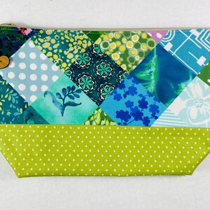 May include: A patchwork zippered pouch with a variety of green and blue patterned fabrics. The pouch features a light green dotted bottom and a wooden heart-shaped tag that reads "Mona Designs". The zipper is gold-toned.