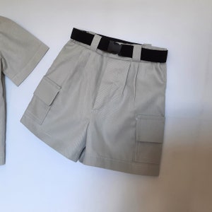 May include: A light tan shorts set with a black belt and buckle. The shorts have two cargo pockets and pleats at the waist. A matching short-sleeved shirt is visible on the left. The set is made of a lightweight fabric.