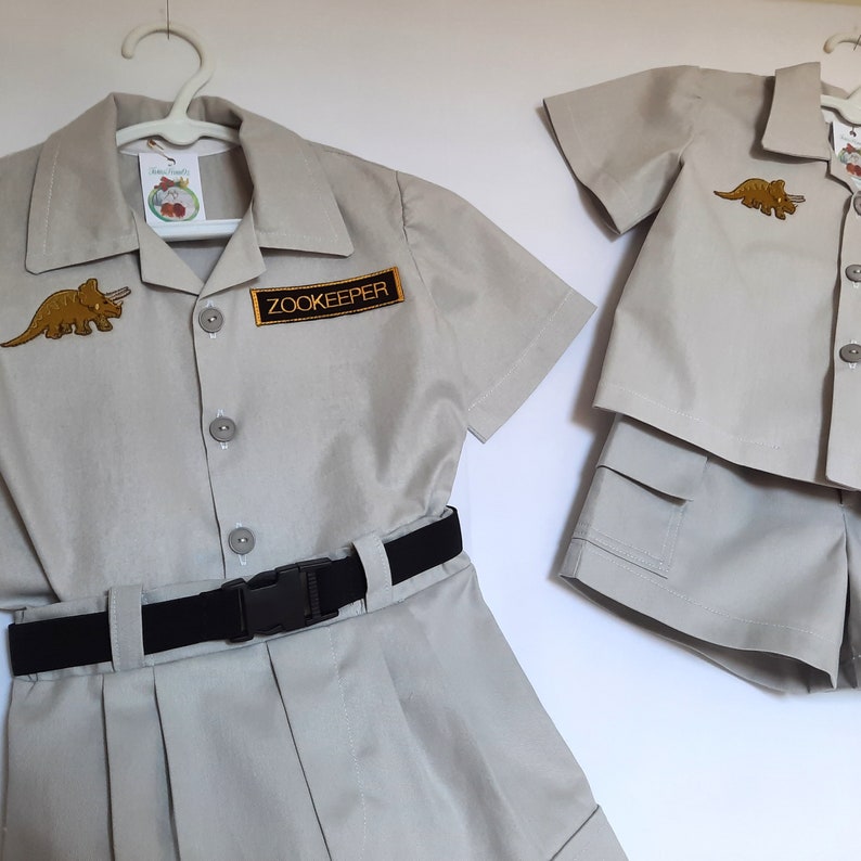 May include: Two khaki-colored zookeeper outfits. One set includes a short-sleeved shirt with a dinosaur patch and a "Zookeeper" patch, a skirt, and a black belt. The other set includes a short-sleeved shirt and shorts, both with a dinosaur patch.