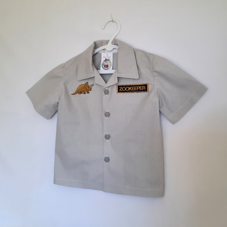 May include: A light gray short-sleeved shirt with a collar, buttons, and a "Zookeeper" patch. An embroidered dinosaur patch is on the left chest. The shirt is hanging on a white hanger.