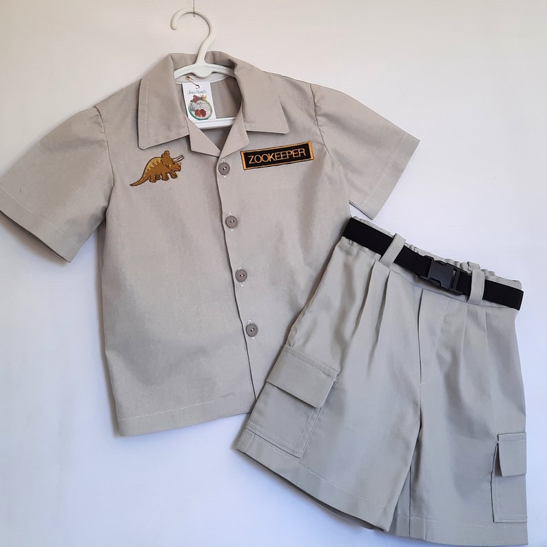 May include: A tan zookeeper costume set, including a short-sleeved button-up shirt with a dinosaur patch and a "Zookeeper" patch, and matching cargo shorts with a black belt. The outfit is displayed on a white hanger.