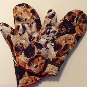 May include: A pair of brown and black oven mitts with a colorful dog print. The mitts have a red scalloped edge and a black label that reads "Je Suis Jackie" with a small heart.