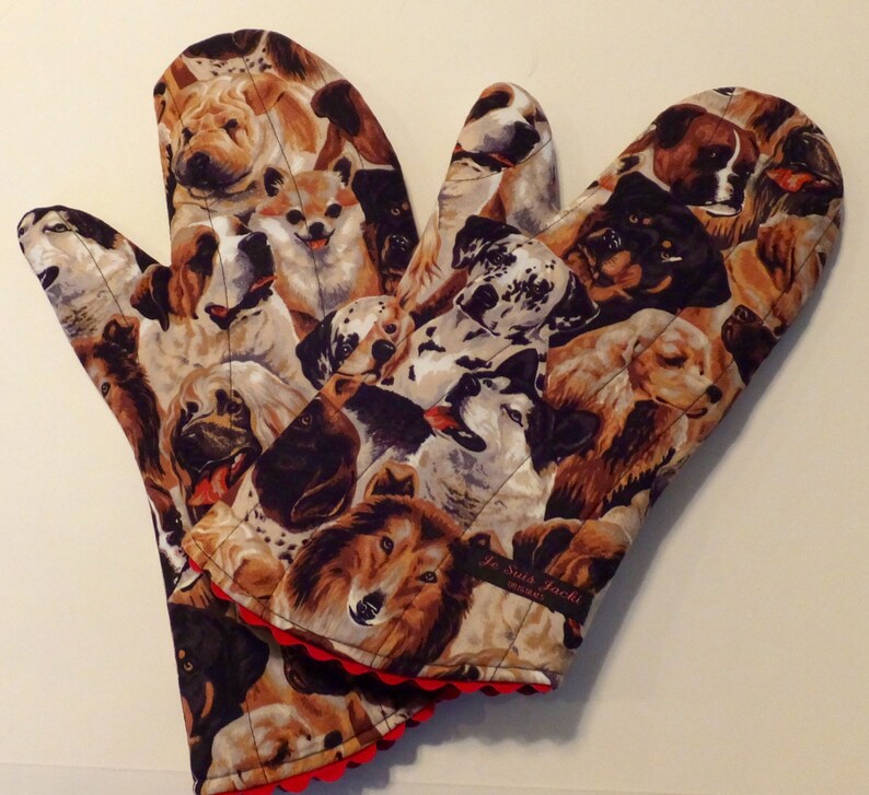May include: A pair of brown and black oven mitts with a colorful dog print. The mitts have a red scalloped edge and a black label that reads "Je Suis Jackie" with a small heart.