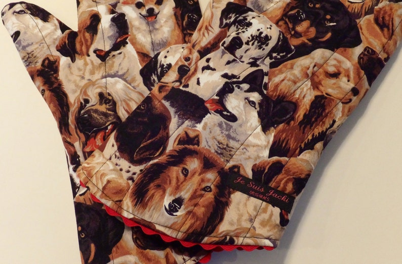 May include: A pair of brown and beige oven mitts with a quilted design featuring a variety of dog breeds. The mitts have a red scalloped edge and a black label that reads "Je Suis Jacki Originals".