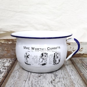 May include: A white enamel chamber pot with a dark blue rim and handle. The front features black printed text reading Mme. Worth's Corsets, New Bond Street, along with three detailed black and white illustrations of figures in Victorian-era clothing.