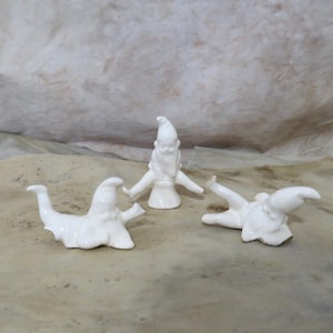May include: Three white ceramic gnome figurines in various poses. One gnome stands upright, arms outstretched, while the other two are lying down. The gnomes have pointed hats and beards.