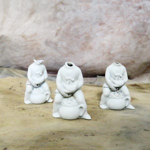 May include: Three small, white ceramic figurines. Each figure is seated with its hands covering its face. The figurines have a bulbous body and are missing their heads. The background is a neutral, textured surface.