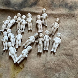 Pu&ograve; includere: Una collezione di piccole bambole antiche in biscuit in varie pose. Le bambole sono bianche con dettagli visibili, sparse su una superficie marrone strutturata. Le bambole sono in diverse posizioni, alcune in piedi e altre sdraiate.