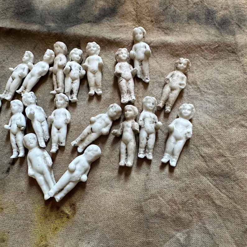 Pu&ograve; includere: Una collezione di piccole bambole antiche in biscuit in varie pose. Le bambole sono bianche con dettagli visibili, sparse su una superficie marrone strutturata. Le bambole sono in diverse posizioni, alcune in piedi e altre sdraiate.