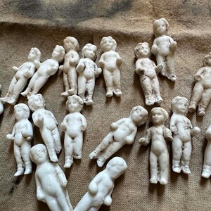 Pu&ograve; includere: Una collezione di piccole bambole antiche in biscuit in varie pose. Le bambole sono bianche con dettagli visibili, alcune con leggera scolorimento. Le bambole sono disposte su una superficie strutturata a tonalit&agrave; neutra.