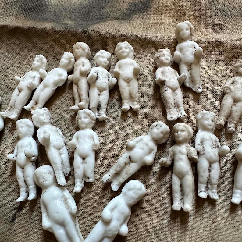 Pu&ograve; includere: Una collezione di piccole bambole antiche in biscuit in varie pose. Le bambole sono bianche con dettagli visibili, alcune con leggera scolorimento. Le bambole sono disposte su una superficie strutturata a tonalit&agrave; neutra.