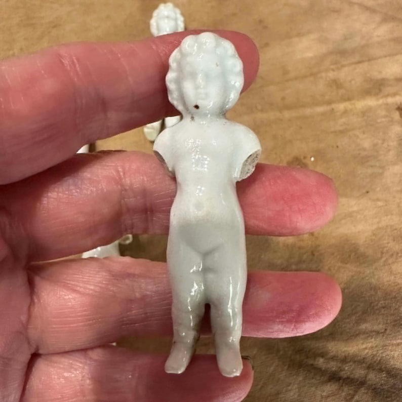 May include: A small, white, unglazed porcelain figurine of a standing child with curly hair. The figure is armless, featuring a smooth, glossy finish and a simple, molded form. It is a miniature doll component, crafted from white ceramic material.