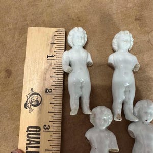 May include: Four small white porcelain doll figurines with molded hair and facial features, positioned next to a wooden ruler showing a scale of approximately 3 inches in height for the standing figures, with visible arm breakage.