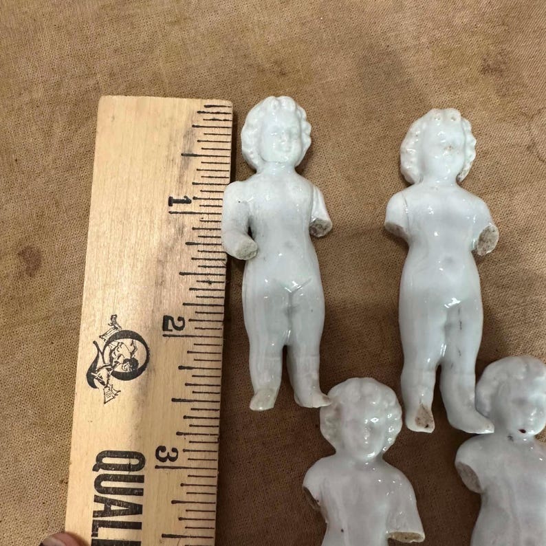May include: Four small white porcelain doll figurines with molded hair and facial features, positioned next to a wooden ruler showing a scale of approximately 3 inches in height for the standing figures, with visible arm breakage.