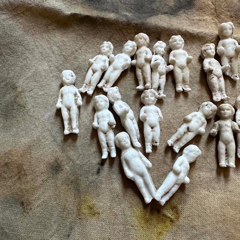 Pu&ograve; includere: Una collezione di piccole statuette di bambole in porcellana bianca. Le bambole sono in varie pose, alcune in piedi e altre sdraiate. Lo sfondo &egrave; un tessuto marrone screziato.