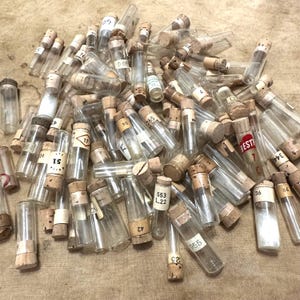 May include: A large collection of small, clear glass vials with cork stoppers. Each vial features a white paper label with printed numbers or text, including labels like 51, 553 L.22, 955, 36, and a red label marked BESTR 7 C. The vials are scattered in a pile.