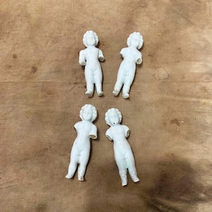 May include: Four small, white, unglazed porcelain doll figures arranged in a group. Each figurine features a molded human form with a defined head, torso, and legs, but lacks arms. The figures have a smooth, matte finish and stand in an upright position.