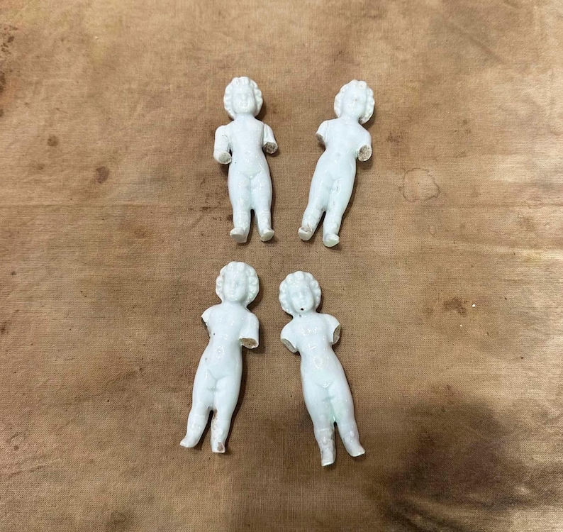 May include: Four small, white, unglazed porcelain doll figures arranged in a group. Each figurine features a molded human form with a defined head, torso, and legs, but lacks arms. The figures have a smooth, matte finish and stand in an upright position.