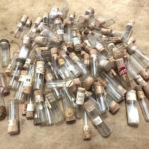 May include: A large collection of small, clear glass vials with cork stoppers. Each vial features a white paper label with printed numbers or text, including labels like 51, 553 L.22, 955, 36, and a red label marked BESTR 7 C. The vials are scattered in a pile.