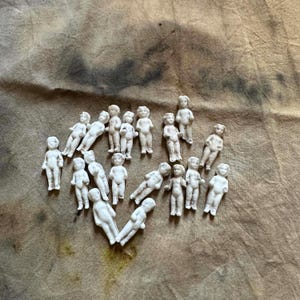 Pu&ograve; includere: Una collezione di piccole statuette di bambole in porcellana bianca disposte su un tessuto strutturato color beige. Le bambole sono in varie pose, alcune in piedi e altre sdraiate. La disposizione generale forma una forma di cuore.