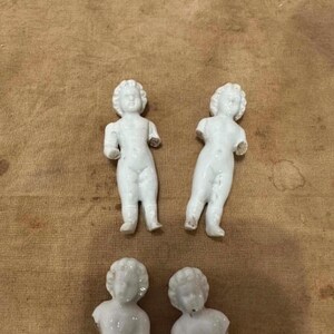 May include: Four white porcelain doll figures with molded hair and facial features. Two full-body figures stand upright with arms extended, while two partial figures show only the head and torso. The figures have a smooth, glossy finish.
