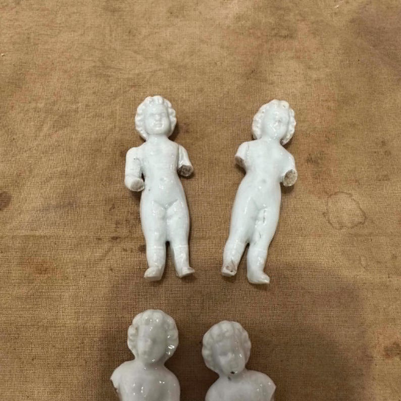 May include: Four white porcelain doll figures with molded hair and facial features. Two full-body figures stand upright with arms extended, while two partial figures show only the head and torso. The figures have a smooth, glossy finish.