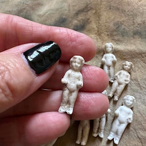 Pu&ograve; includere: Una collezione di piccole bambole antiche in biscuit. Le bambole sono bianche con dettagli visibili e sono in varie pose. Le bambole sono alte circa 4 cm. Lo sfondo &egrave; un tessuto strutturato color beige.