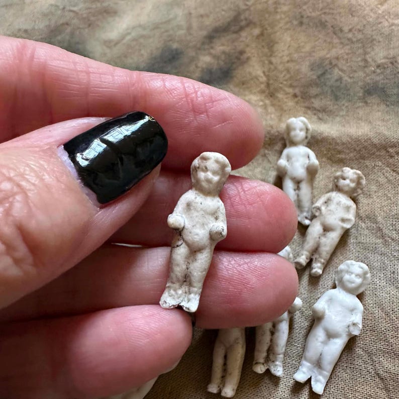 Pu&ograve; includere: Una collezione di piccole bambole antiche in biscuit. Le bambole sono bianche con dettagli visibili e sono in varie pose. Le bambole sono alte circa 4 cm. Lo sfondo &egrave; un tessuto strutturato color beige.