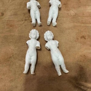 May include: Four small, white, unglazed porcelain doll figures arranged in two rows. Each figure features a molded human form with a defined head, torso, and legs, but lacks arms. The surface of the porcelain is smooth and matte, showing a uniform white color across all four pieces.