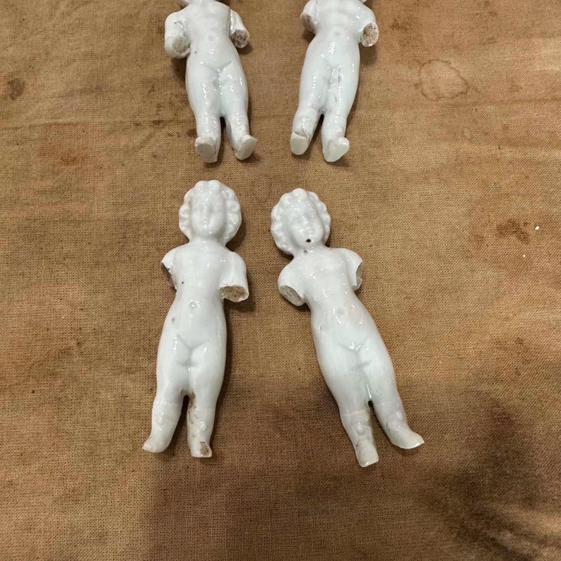 May include: Four small, white, unglazed porcelain doll figures arranged in two rows. Each figure features a molded human form with a defined head, torso, and legs, but lacks arms. The surface of the porcelain is smooth and matte, showing a uniform white color across all four pieces.