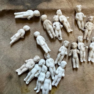 Pu&ograve; includere: Una collezione di piccole statuette di bambole in porcellana bianca sparse su una superficie strutturata e marrone. Le bambole sono in varie pose, alcune in piedi e altre sdraiate. Le bambole hanno lineamenti dettagliati e sono in stile vintage.