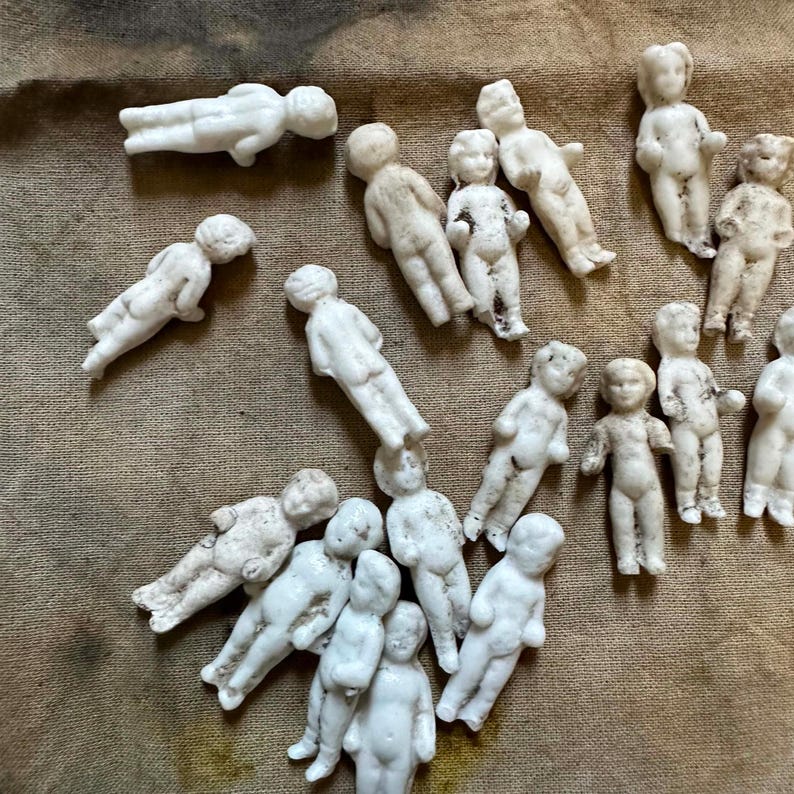 Pu&ograve; includere: Una collezione di piccole statuette di bambole in porcellana bianca sparse su una superficie strutturata e marrone. Le bambole sono in varie pose, alcune in piedi e altre sdraiate. Le bambole hanno lineamenti dettagliati e sono in stile vintage.