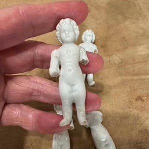 May include: A collection of small, white, unglazed porcelain frozen Charlotte dolls. Each doll features a molded, detailed face with curly hair and a smooth, standing body. The dolls are depicted in a nude, upright position with arms at their sides.