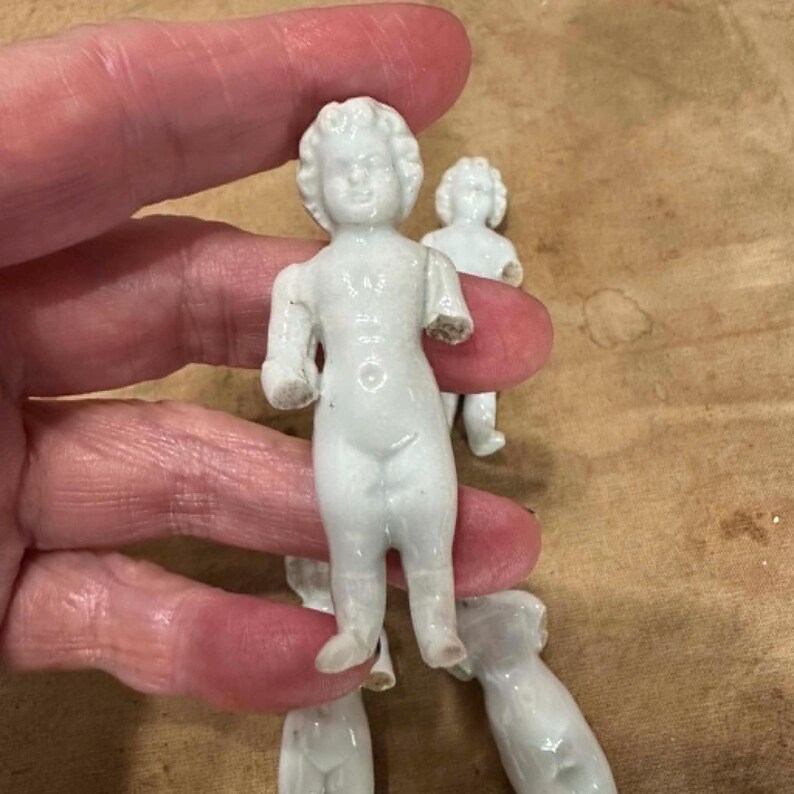 May include: A collection of small, white, unglazed porcelain frozen Charlotte dolls. Each doll features a molded, detailed face with curly hair and a smooth, standing body. The dolls are depicted in a nude, upright position with arms at their sides.