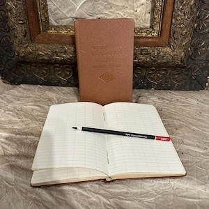 May include: An open, lined notebook with a black pencil on top. A closed brown notebook is behind it. The open notebook has a grid pattern and the pencil has the text "PEPY Charcoal Selection SOFT".