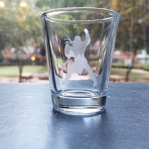 May include: A clear shot glass with a warrior figure etched on the front. The glass is cylindrical with a thick base and is sitting on a dark surface. The background shows a blurred outdoor scene.