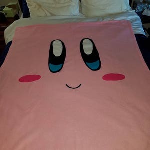 Könnte beinhalten: Eine rosa Fleece-Decke mit einem Comic-Gesicht eines rosa Charakters mit schwarzen Augen, blauen Pupillen und einem schwarzen Lächeln. Der Charakter hat rote Wangen.