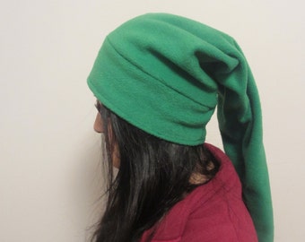 May include: A green fleece hat with a long pointed tail. The hat is being worn by a person with long dark hair and a red shirt.